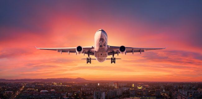 Airplane is flying in colorful sky over city at sunset. Landscape with passenger airplane, skyline, purple sky with red and pink clouds at dusk. Aircraft is landing at twilight. Aerial view of plane